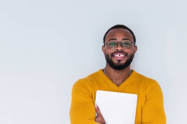 Pozitif sakallı Afro-Amerikan erkek, sarı kazaklı ve gözlüklü, elinde not defteri, kameraya bakıyor ve beyaz arka planda duruyor.