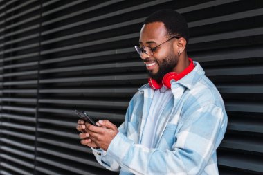 Cep telefonlu mutlu Afrikalı erkek modern binanın duvarının yanında dururken kablosuz kulaklıkla müzik dinliyor.