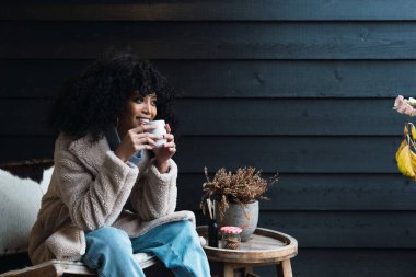 Pozitif Afro-Amerikalı, kıvırcık siyah saçlı, kalın paltolu, bardaktan sıcak içecek içen, evin dışında oturan ve başka tarafa bakan genç bir kadın.