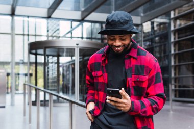 Şapkalı ve kareli tişörtlü Afro-Amerikan adam cam duvarlı ve cep telefonlu bir binanın lobisinde yürüyor.