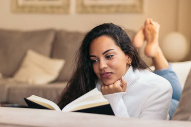 Evde kitap okuyan genç bir kadın. Sakin ve sakin bir tavırla, boş zamanlarının tadını çıkarıyor.