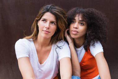 Dingin, düşünceli, çok ırklı bayan arkadaşlar. Kahverengi arka plana bakarken kulaklıklardan şarkı dinliyorlar.