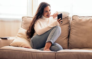 Evinde rahat bir kanepede oturan genç bir kadın, cep telefonunu kullanıyor. Kadın bir dakikalık dinlenmenin ve cep telefonuyla bağlantının keyfini çıkarıyor.