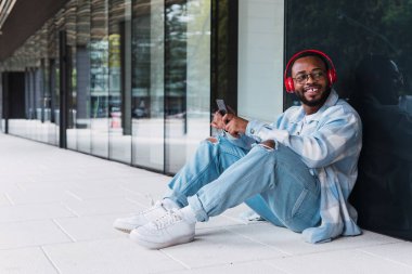 Modern binanın duvarına bakarken cep telefonuyla kablosuz kulaklıkla müzik dinleyen mutlu Afrikalı Amerikalı bir erkek.