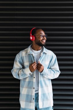 Şık zevkli Afro-Amerikan erkek şık ceketli kırmızı kablosuz kulaklıklarla müzik dinlerken siyah duvarın yanındaki sokağa bakıyor.