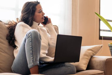Evdeki kanepesinde oturan genç bir kadın, akıllı bir telefonla konuşurken dizüstü bilgisayarıyla çalışıyor..
