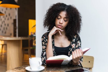 Afro saçlı genç bir kadın kafeterya masasına yaslanmış ders kitabı okurken.