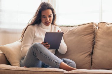 Genç bir kadın evinde bir kanepede oturuyor, tablet kullanıyor..