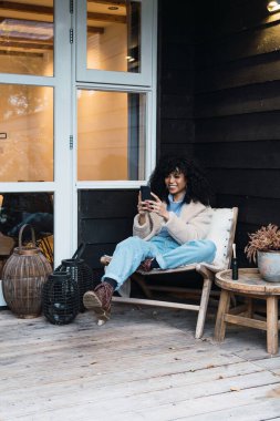Genç, gülümseyen, kıvırcık, siyah saçlı, şık, sıcak elbiseli, terasta cep telefonuyla koltukta dinlenen bir kadın.