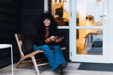 Kot pantolon ve sıcak ceket giyen, terasta sandalyeye oturup gözlerini kaçıran neşeli, genç Afrikalı Amerikalı bir kadın.