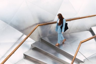 Yukarıdan bakıldığında, uzun saçlı, zarif giyimli, el çantalı, yüzü olmayan, geniş bir binanın koridorunda yürüyen kadın görünüyor.