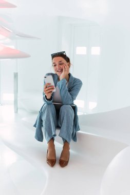 Zarif kadın tüm vücudu beyaz duvarlı, aydınlık modern odada otururken cep telefonuyla sosyal medyayı kaydırıyor.