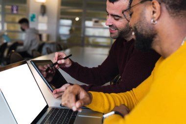 Sırıtkan çok ırklı sakallı ortaklar birlikte çalışma merkezinde dizüstü bilgisayar ve tablet üzerinde çalışıyorlar.