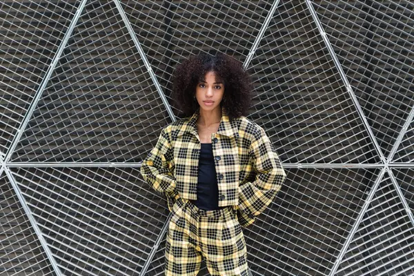 Young ethnic female in trendy checkered suit with hands behind back looking at camera in daytime