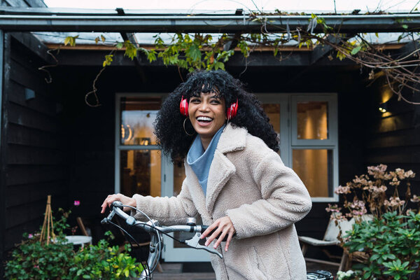 Side view smiling young African American female with curly dark hair wearing warm coat walking with bicycle while listening to music in headphones near house in autumn day