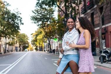 İki genç kadın, arkadaşlar, yaya geçidinden geçiyorlar. Gülerek ve kollarını tutarak yürürler.