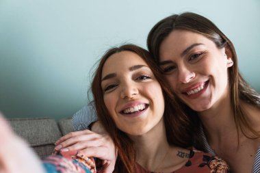 Genç ve mutlu bayan arkadaşlar günlük kıyafetler giyip kucaklaşıyorlar ve cep telefonuyla açık oturma odasında selfie çekiyorlar.