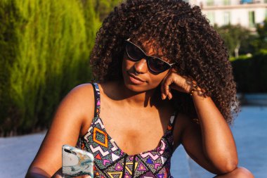 Moda kıyafetli ve güneş gözlüklü genç siyahi kadın sınırda bacak bacak üstüne atarak oturuyor ve yaz günü akıllı telefonlara bakıyor.