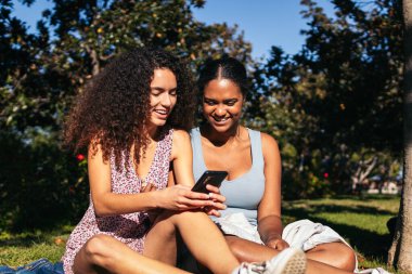 Yazın açık havada akıllı telefon kullanan afro saçlı iki kız. Onlar arkadaşlar, şehir parkındaki bir toplantıda iyi vakit geçiriyorlar..