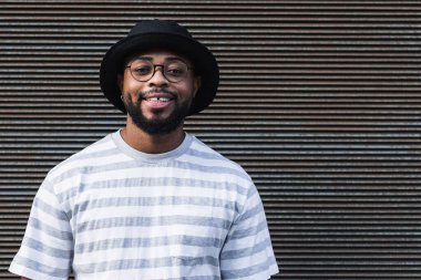 Gözlüklü, gözlüklü, modaya uygun kıyafet ve şapka takan parlak bir şekilde gülümseyen ve kapalı duvara bakarken kameraya bakan mutlu Afrikalı Amerikalı sakallı adam.