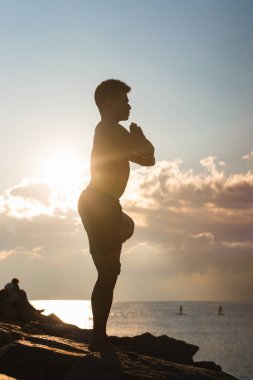 Side view silhouette of unrecognizable male balancing on Tree pose with arms up while practicing yoga against cloudy sundown sky clipart