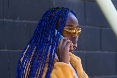 Mavi örgülü, sarı elbiseli, şık siyah bir kadın sokakta akıllı telefondan konuşuyor.