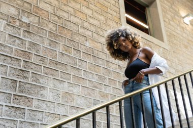 Düşük açılı, yırtık kot pantolon giyen, merdivenlere yaslanmış ve başka yöne bakan, kendine güvenen Afro-Amerikan kadın.