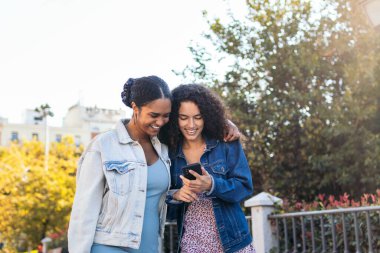 İki Latin Amerikalı arkadaş sokakta yürüyor, eğleniyor ve akıllı telefon kullanarak gülüyorlar..