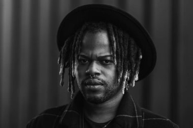 Gayri resmi sakallı, şapkalı, sokakta duran kameraya bakan etnik bir adam.