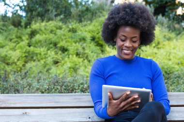 Parkta bankta oturan ve yeni başlatılan projede çalışırken tablet kullanan içerikli Afrikalı Amerikalı kadın serbest yazar