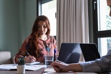 İçerik çalışanları ya da öğrenciler gündelik kıyafetlerini giyer ve modern iş yerinde masa başında otururken not defterine not alır.