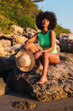 Kıvırcık saçlı, mayo giymiş, hasır şapkalı ve yazın günbatımında kameraya bakan sakin Afro-Amerikan kadın.