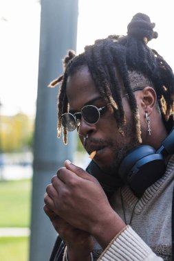 Ekmekli, güneş gözlüklü, kulaklıklı ve sigara yakan havalı Afrikalı Amerikalı adam.