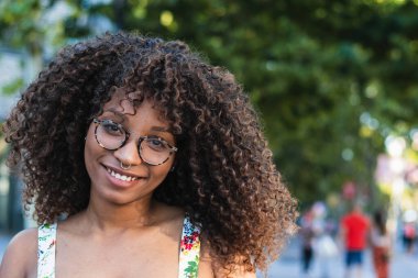 Kıvırcık saçlı mutlu zenci kadın yaz günü parkta kameraya bakıp gülümsüyor.