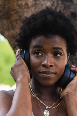 Mutlu Afrikalı Amerikalı kadın ağacın yanında dururken kablosuz kulaklıkla müzik dinliyor.