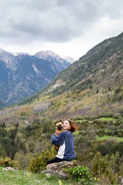 Pireneler 'deki dağ sırasındaki macera sırasında kayanın üzerinde oturmuş Fransız Bulldog' unu kucaklayan kadın yürüyüşçünün yan görüntüsü kameraya bakıyor.