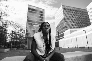 Sınırda oturmuş, caddede kameraya bakan, modaya uygun giyinmiş, duygusuz, genç bir kadın.