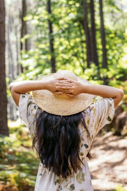 Arka planda, ormanda kolları başının üzerinde hasır şapka giyen bir kadın görülüyor. Doğal ortamda doğal tatil ve rahatlama kavramı