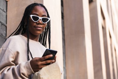 Gri tuğla duvara yaslanan ve şehir caddesinde akıllı telefonlara bakan gündelik kıyafetli genç bir etnik kadın.