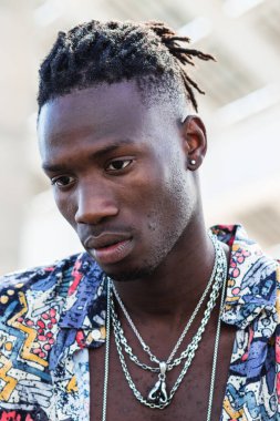 Ciddi Afro-Amerikan erkek, renkli gömlekli ve kot pantolonlu, taş basamaklarda oturuyor ve şehir sokağına bakıyor.