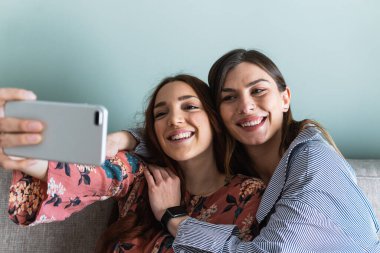 Genç ve mutlu bayan arkadaşlar günlük kıyafetler giyip kucaklaşıyorlar ve cep telefonuyla açık oturma odasında selfie çekiyorlar.