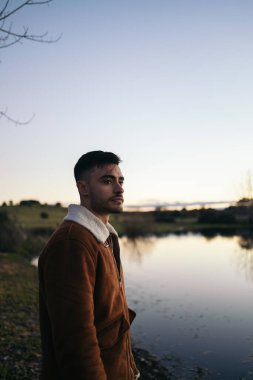 Kırsaldaki bir gölde gün batımını izleyen sakallı çekici genç bir adam. Sakin görünüyor. Doğadaki güzel manzaranın tadını çıkarıyor.
