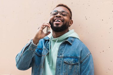 Yetişkin hippi sakallı Afro-Amerikan erkek sıradan kıyafetler ve gözlüklerle telefonla konuşuyor ve haberlerle tatmin olurken sokakta bej taştan duvarın önünde dikiliyor.