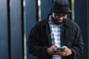 Neşeli yetişkin sakallı Afro-Amerikan erkek. Günlük kıyafet ve şapka giymiş. Duvarın karşısında duruyor ve akıllı telefondan mesaj yolluyor.
