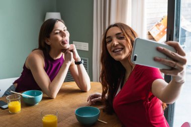 Neşeli genç bayan arkadaşlar gündelik giysiler içinde birlikte lezzetli bir öğle yemeği yiyorlar ve modern bir dairede yemek masasında oturup sohbet ederken akıllı telefon kullanıyorlar.