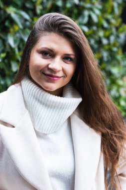 Parktaki yeşil çalıların yanında dinlenirken beyaz süveterli, zarif, uzun saçlı esmer, hoş bir adam.