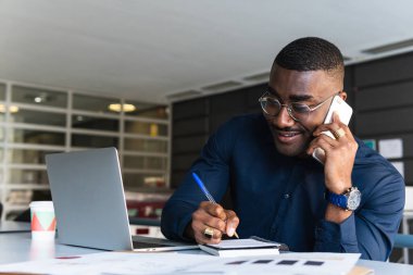 Gözlüklü gülümseyen Afrikalı Amerikalı erkek ve masada resmi giysiler cep telefonuyla konuşuyor ve not defterine, netbook 'un yanında not defteri ve belgeyle hafif iş yerinde içeceklerle birlikte not alıyor.