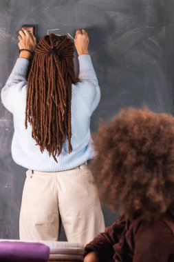 Rastalı kadın öğretmenin sınıfta masasında oturan siyah kız için tahtaya matematiksel örnekler yazması.