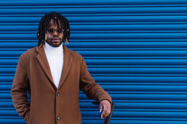 Afro örgülü, şık bir ceket ve güneş gözlüğü takan, kendine güvenen Afro-Amerikan hippi erkek. Mavi çizgili duvara karşı elektrikli scooter 'ın yanında duruyor.