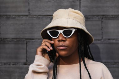 Gri tuğladan duvara yaslanan ve şehir caddesinde akıllı telefon konuşan gündelik kıyafetli genç bir etnik kadın.